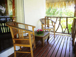 A small porch with wooden chairs and a table, offering a view of tropical plants in a peaceful outdoor setting.