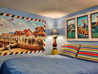 A bedroom with coastal-themed decor, including a bed with striped pillows and a blue quilt, a painting of a boardwalk with people, and a seascape artwork above the bed, all illuminated by a lamp.