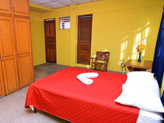 A bedroom with yellow walls features a double bed with a red quilt, white pillows, and towels shaped into a heart. There is wooden furniture, a small table with flowers, and a wardrobe.