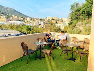 Two people sit at a table with coffee on a balcony overlooking a cityscape and hills.