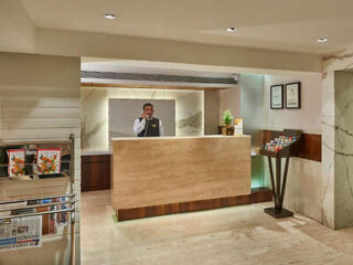 Hotel reception area with a front desk attended by a staff member on the phone. The area includes a magazine rack, a display of brochures, and framed certificates on the wall.