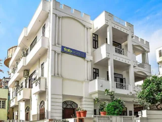 Three-story white building with balconies and a sign reading "fab hotels" on the second floor. The structure has a clear, sunny sky in the background.