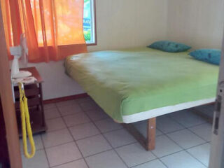 A simple bedroom with a green bedspread, a window with orange curtains, and a small wooden nightstand.