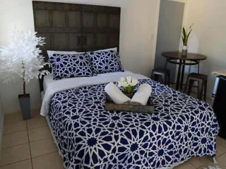 A neatly made bed with a blue and white geometric patterned comforter. A tray with folded towels and flowers is placed on the bed, and there is a small round table with stools in the corner.