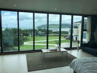 A room with floor-to-ceiling windows overlooks a grassy area, trees, and the ocean. A coffee table stands on a rug in front of the windows, and part of a bed is visible in the foreground.