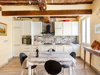 A kitchen with white cabinetry, patterned backsplash, and wooden beams. A table with a tablecloth, wine bottle, and glasses is centered, accompanied by four black chairs. A window allows natural light.