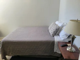 A neatly made bed with a gray quilt, two pillows, a bedside table with a lamp, a remote control, and a small book in a minimally decorated room.