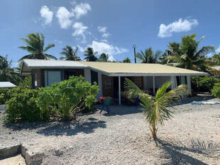 A beachside house with a wide porch, surrounded by palm trees and tropical plants, set on a gravel path.