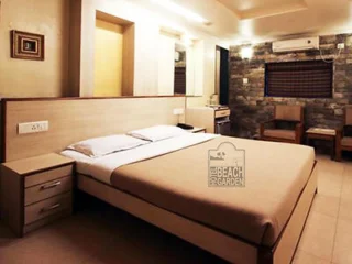 Hotel room with a double bed, bedside tables, a wall-mounted air conditioner, a small fridge, a chair, and a stone-textured wall. Sign reads "Hotel Beach Garden".