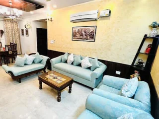 Living room with light blue sofas, a wooden coffee table, wall-mounted air conditioner, framed picture, and corner shelf. Dining area is visible in the background. Wall decor includes a clock.