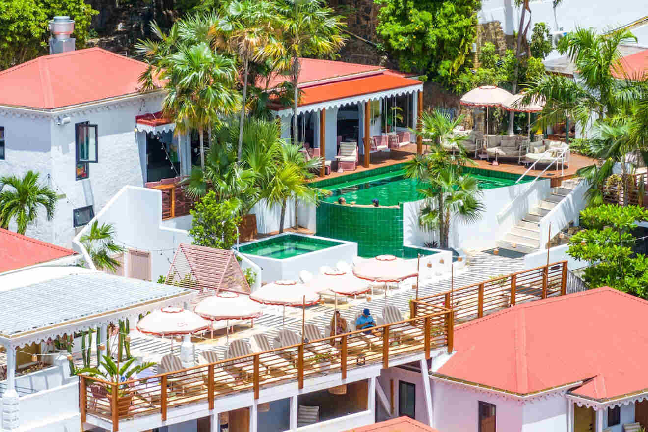 Aerial view of a resort featuring white buildings with red roofs, outdoor seating under umbrellas, a tiered pool, and palm trees.