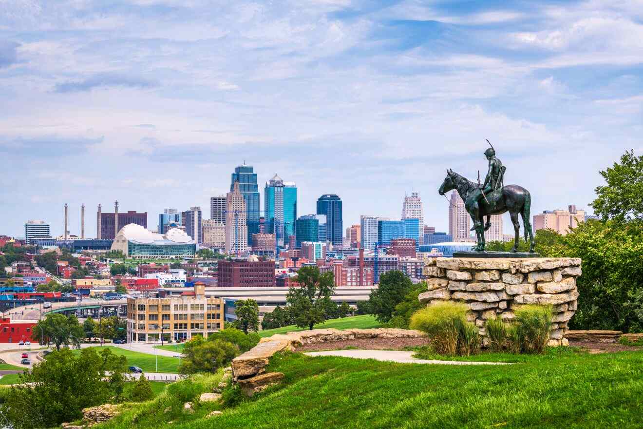 A statue of a Native American on horseback overlooking the Kansas City skyline, with modern skyscrapers and landmarks like the Kauffman Center visible in the distance.