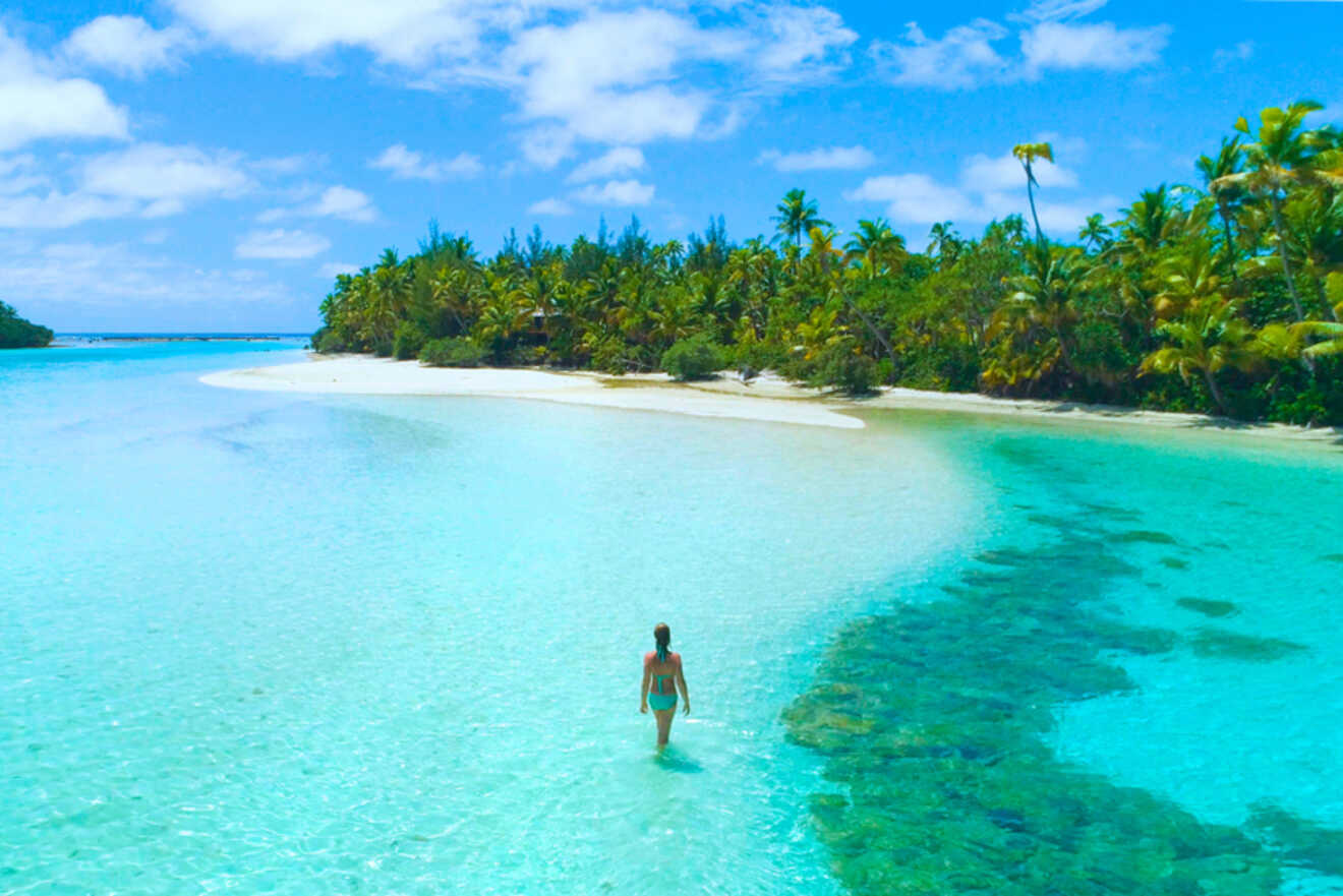 Woman standing in shallow, crystal-clear water on a sandy beach surrounded by tropical palms in the Cook Islands.