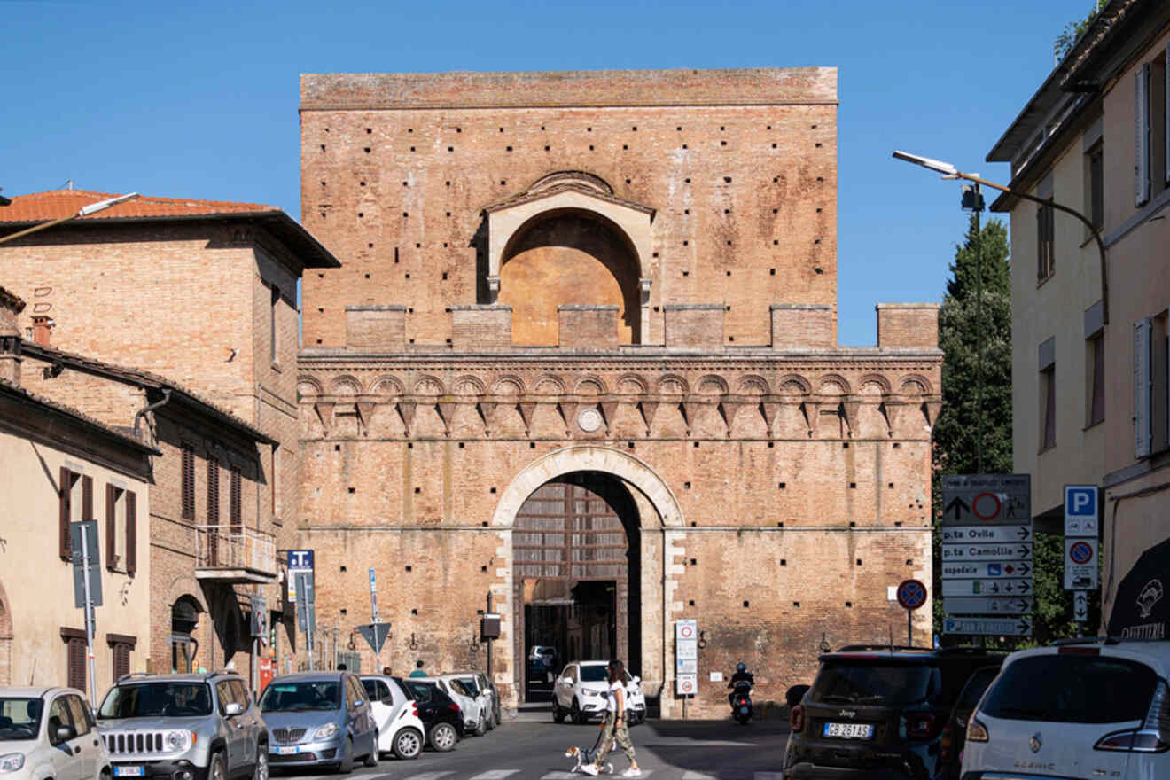 A busy street with cars and pedestrians in front of a large, historic brick gate structure with an arched entrance and small windows. The gate is surrounded by buildings and traffic signs.