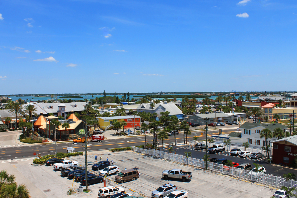 A coastal town is shown with a mixture of buildings, a main street, parked vehicles, and a body of water in the background under a clear blue sky.