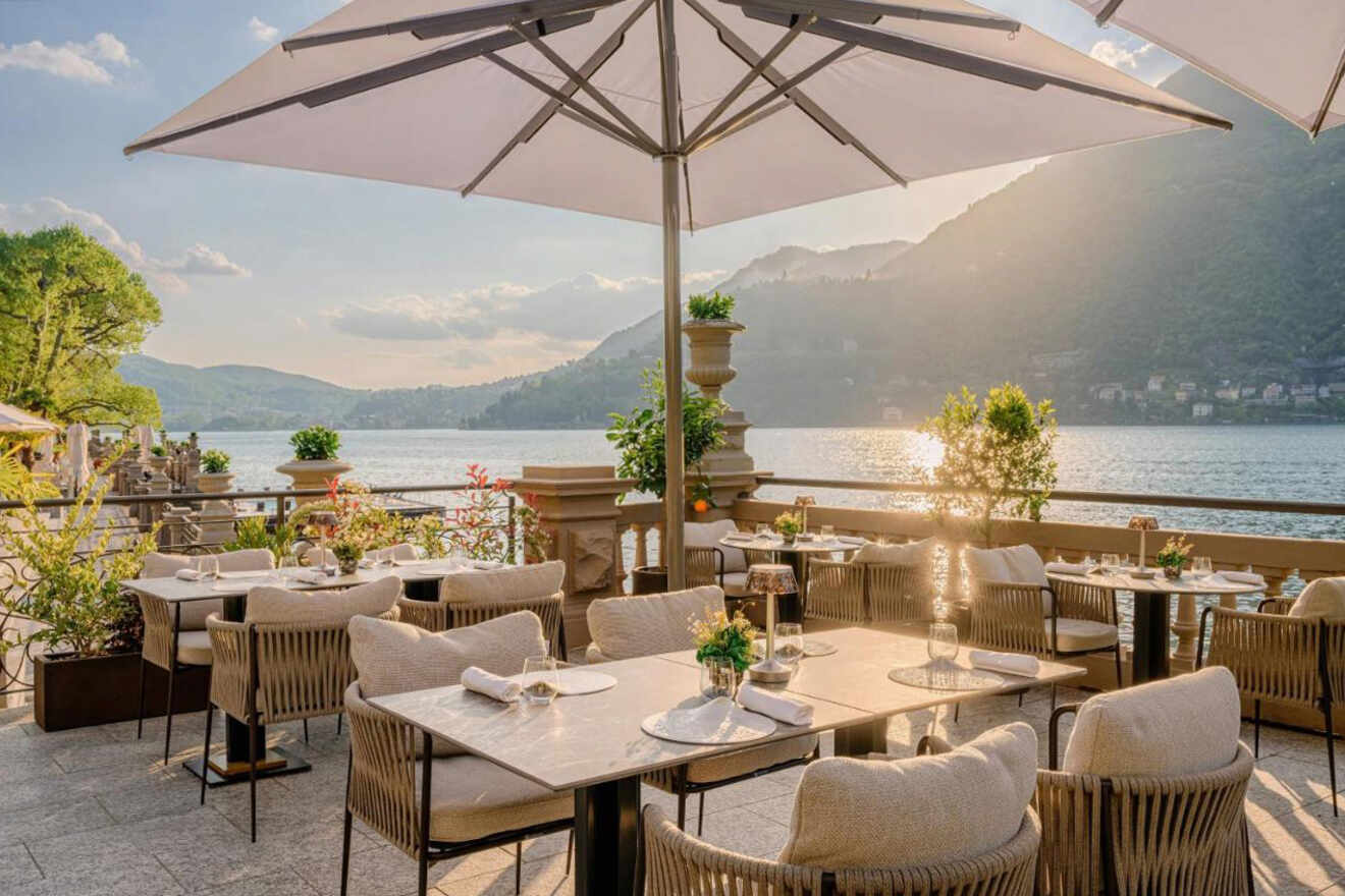 Outdoor restaurant with tables, chairs, and umbrellas overlooking a scenic lake surrounded by mountains. The area is set for dining with table settings in place, and the sun is setting in the background.