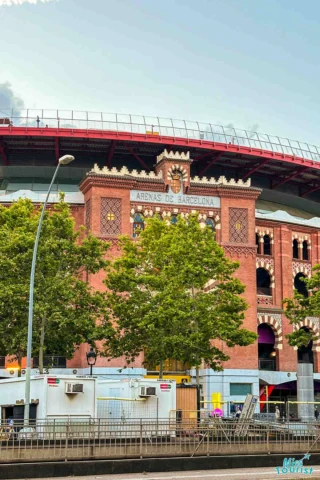 A red-brick building in front of trees, with a sign reading "Arenas de Barcelona" and a curved rooftop visible behind it. Several air conditioning units and construction equipment are in the foreground.