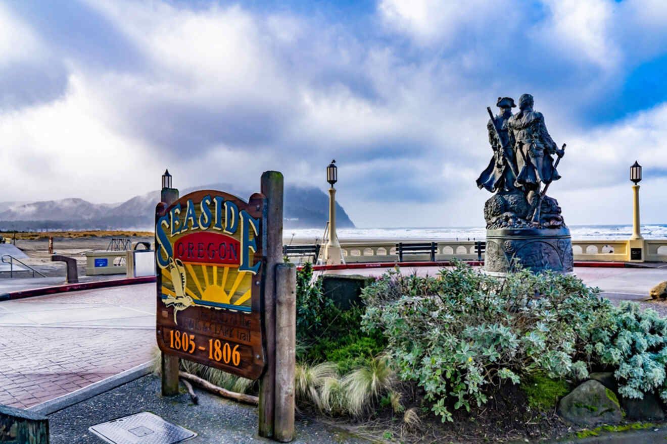 A seaside view in Oregon featuring a "Seaside Oregon" sign, a sculpture of two historical figures, and a cloudy sky with mountains in the background.