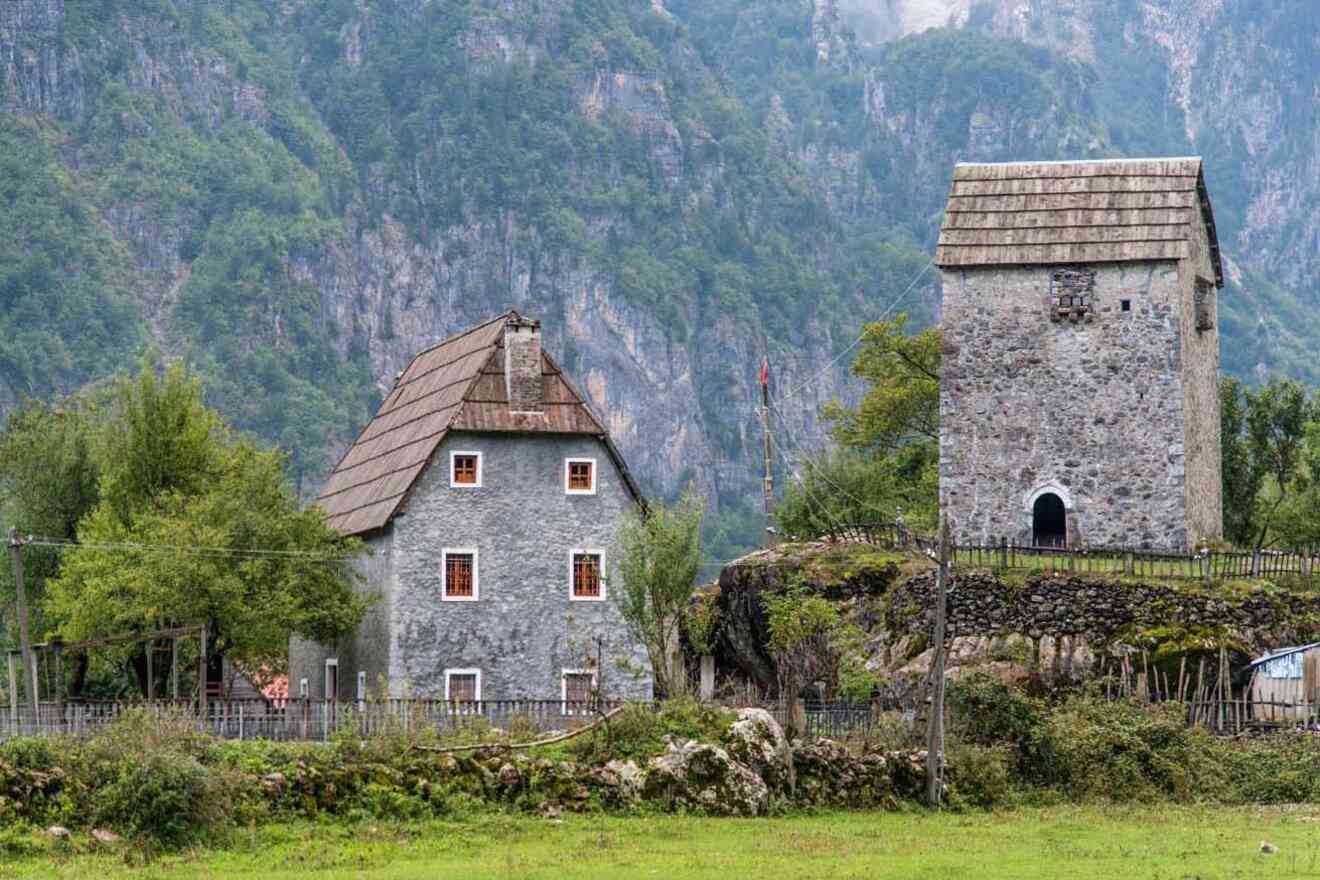 Two historic stone buildings with sloped roofs set against a backdrop of lush green mountains and trees, surrounded by a wooden fence in a rural area.