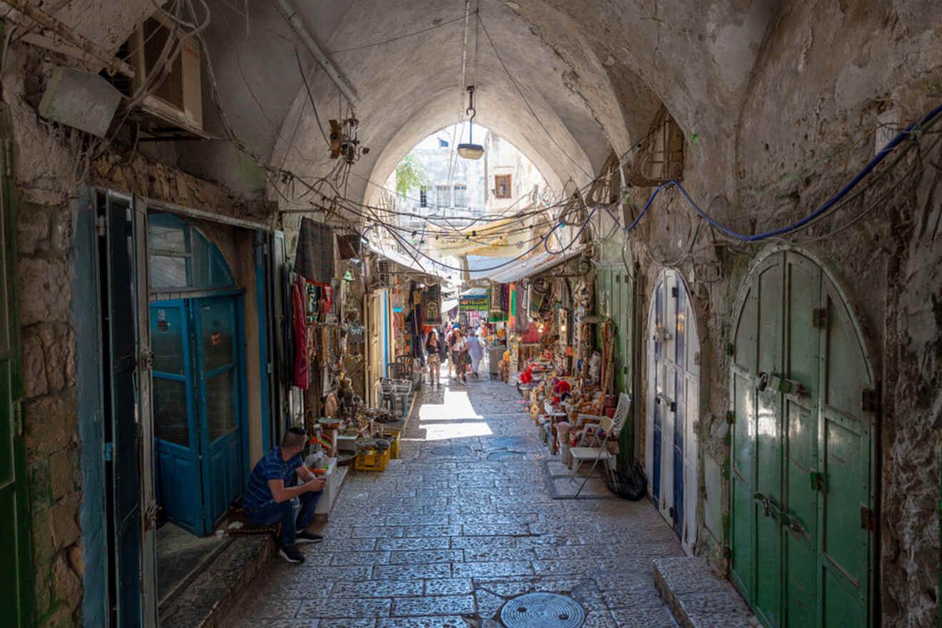 A narrow cobblestone alley with arched ceilings, lined with small shops displaying goods. A few people are walking and shopping in the background. Sunlight filters through the archways.