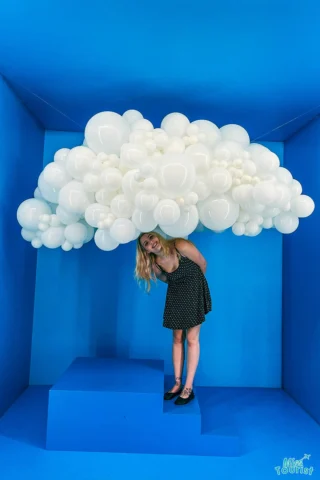 Kathryn, team member of Misstourist, wearing a black polka dot dress standing under a large cluster of white balloons shaped like a cloud, in a blue room.