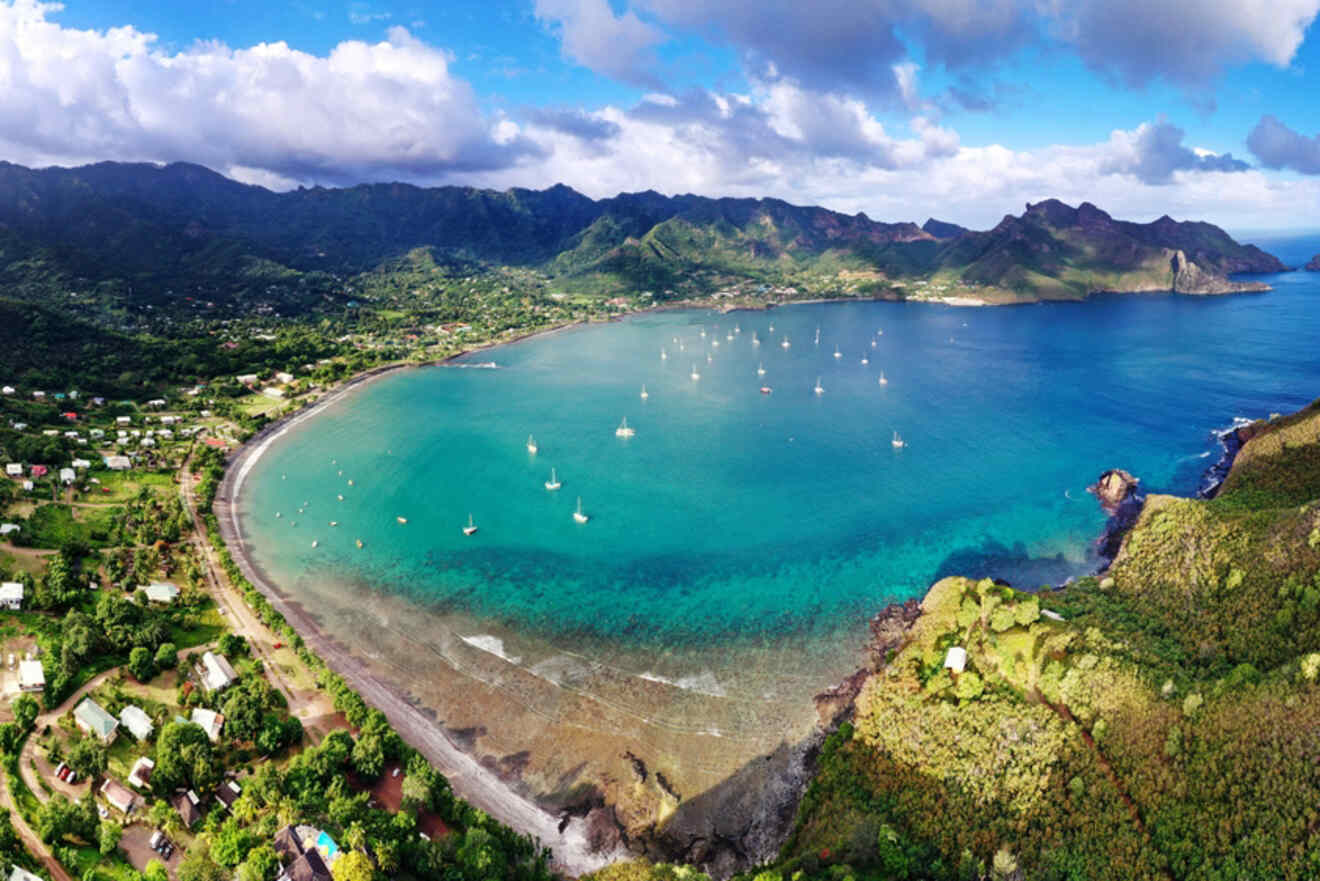 A panoramic view of Nuku Hiva's bay, surrounded by green hills and mountains, with boats dotting the calm turquoise waters and small villages along the coastline.