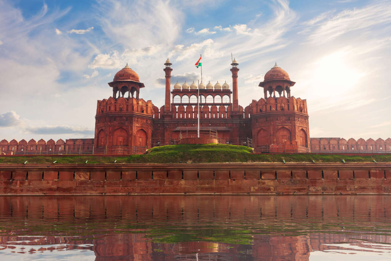 The Red Fort in Old Delhi, a historic red sandstone fortification, reflected in the water under a partly cloudy sky.