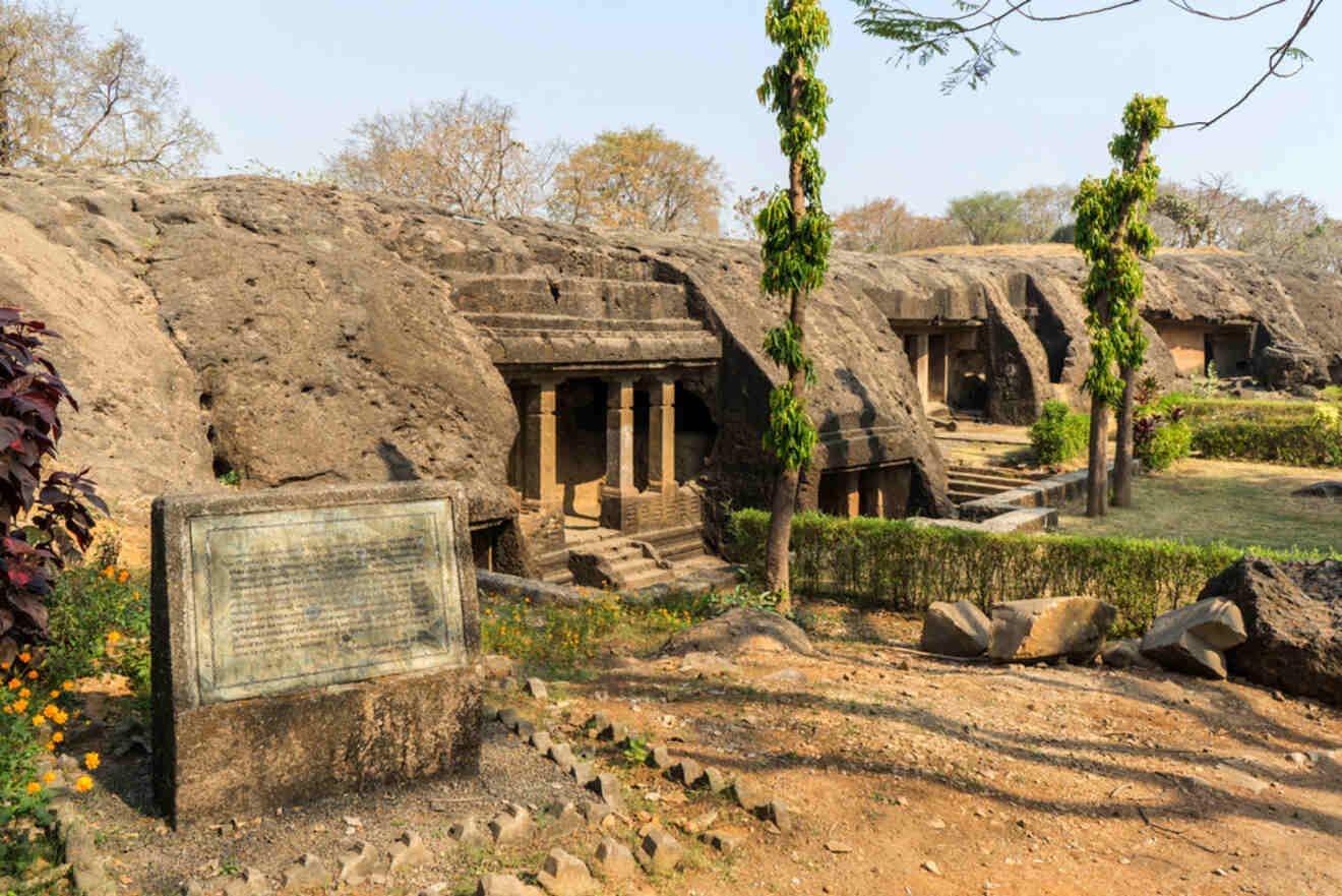 Rock-cut caves with columns and carved entrances, surrounded by greenery and a stone plaque, seen in a historical archaeological site.