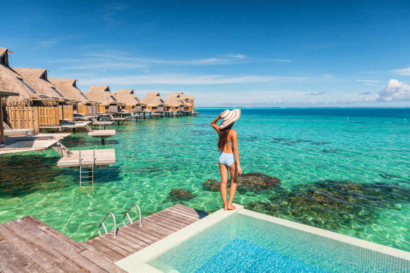 A woman in a swimsuit and sun hat stands beside a pool, overlooking overwater bungalows and clear turquoise ocean water under a clear blue sky.