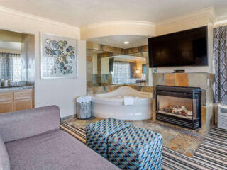 Hotel room with a seating area, a jacuzzi tub, a fireplace, a large flat-screen TV, and modern decor elements. The room has patterned furniture, a striped carpet, and mirrored walls near the tub.