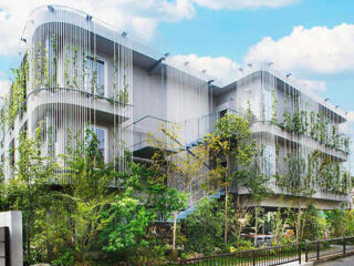 A modern multi-story building with metal accents and greenery growing on the exterior, surrounded by trees and foliage under a partly cloudy sky.