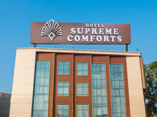 A multi-story building with a sign reading "Hotel Supreme Comforts" on the rooftop, featuring large windows and a symmetrical design against a clear blue sky.