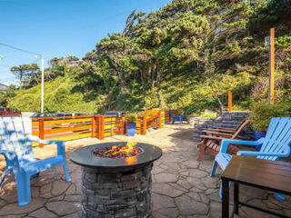 Patio area with a fire pit, surrounded by chairs and tables, set against a backdrop of lush, green trees and clear blue sky.