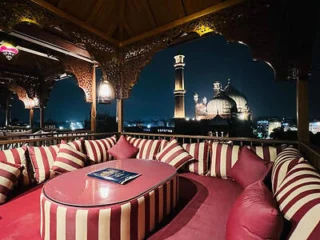 A rooftop lounge with striped cushions and a view of the Jama Masjid lit up at night in the background.