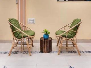 Two rattan chairs with green cushions face each other, with a small circular table holding a blue potted plant between them. The setting is indoors with a tiled floor and a beige wall background.