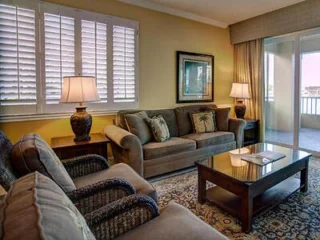 A cozy living room with a sofa, two armchairs, a coffee table, and lamps on side tables. The room features large windows with shutters and a sliding glass door leading to a balcony.