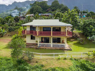 A two-story yellow house with red railings, set against a lush green landscape with mountains in the background.