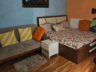 A bedroom with a double bed featuring a colorful patterned duvet and multiple pillows, a gray sofa with yellow-striped cushions, a small white bedside table, and wooden flooring.