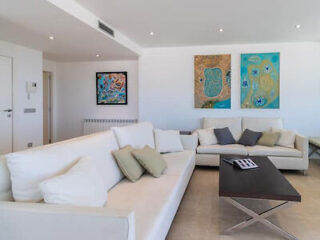 A modern living room with white sofas, a low wooden coffee table, and three abstract art pieces on the wall. The room is well-lit with recessed ceiling lights.