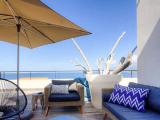 A modern outdoor seating area with a large umbrella, wicker furniture, and views of the ocean