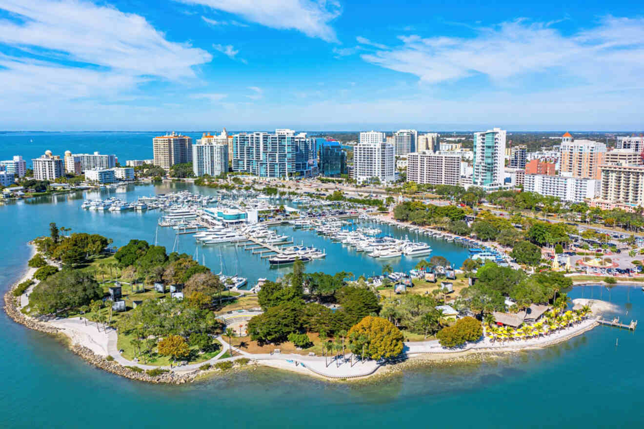 An aerial view of a marina in Sarasota, with luxury yachts docked in the harbor, surrounded by a waterfront park, and framed by a skyline of tall buildings under a sunny blue sky.