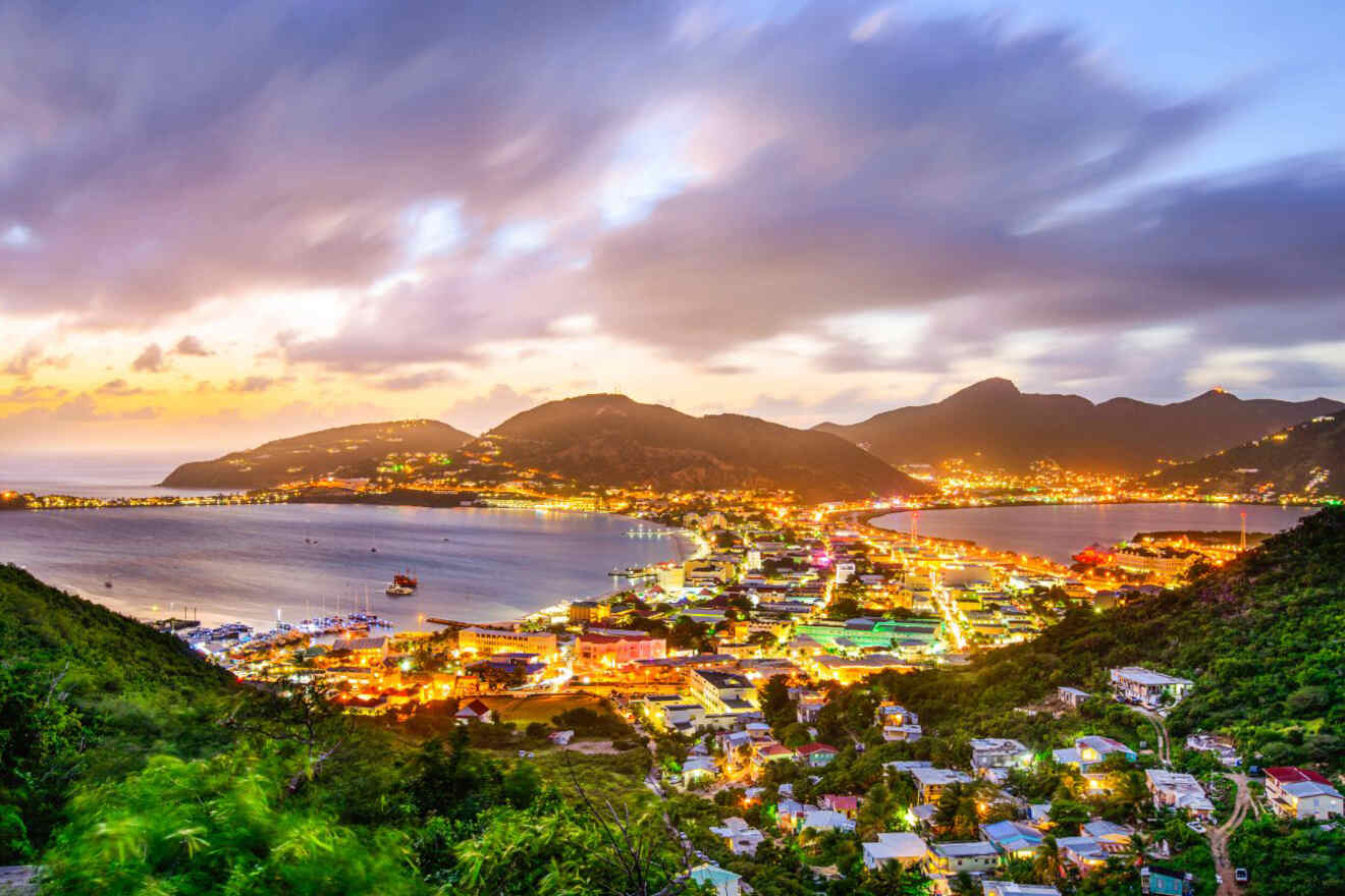 A stunning nighttime view of a coastal town with twinkling lights, curving around two bays, with mountains in the background under a colorful sky.