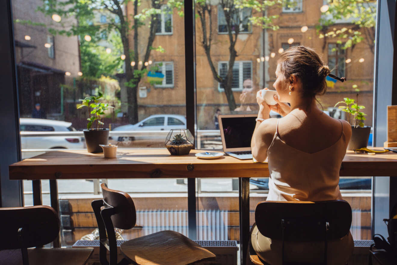 A woman sits at a café window counter, drinking from a cup and using a laptop, with potted plants and sunlight illuminating the interior.