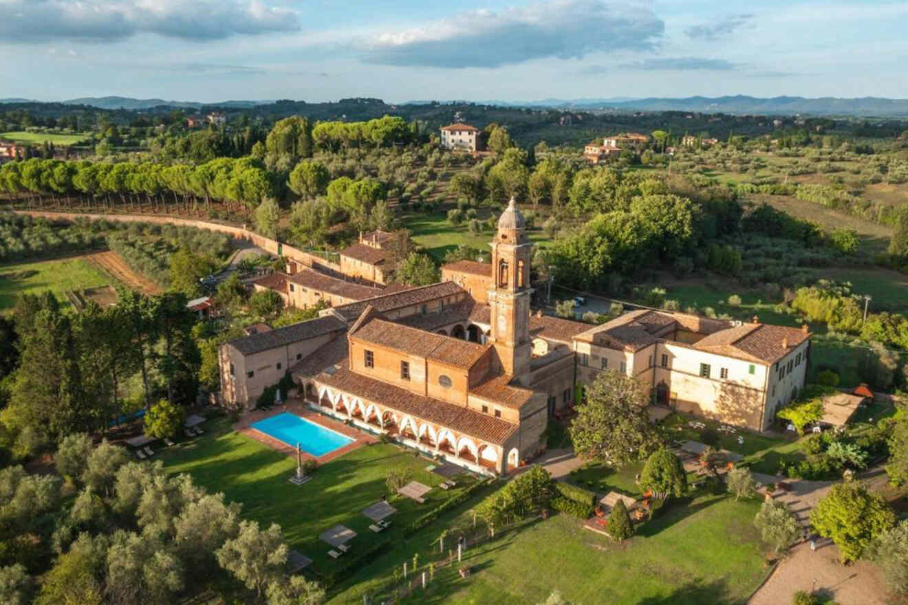 Aerial view of a historic estate with a central bell tower, surrounding buildings, a swimming pool, and lush gardens, set among rolling hills and olive groves.