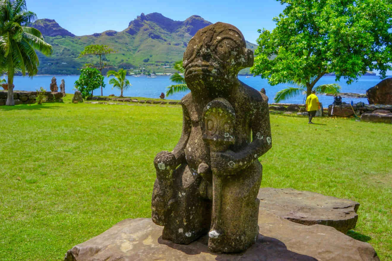 Stone statue of a figure in a grassy area with a mountainous landscape and a body of water in the background. Palm trees and a person in a yellow shirt are visible.