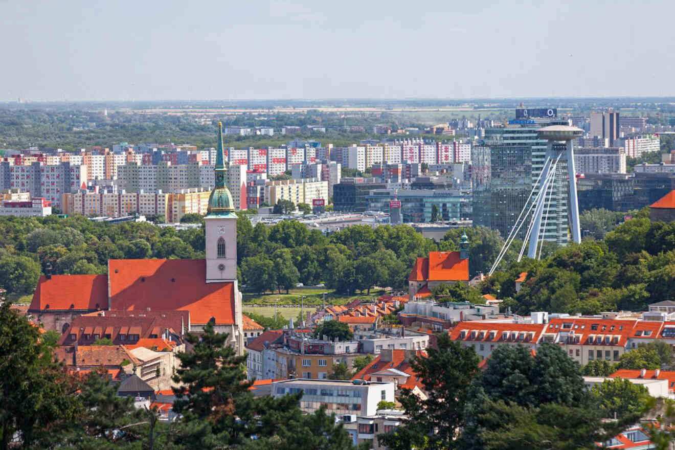 A cityscape view featuring red-roofed buildings, a church with a green spire, lush green trees, and a modern tower with a UFO-like structure near the top.