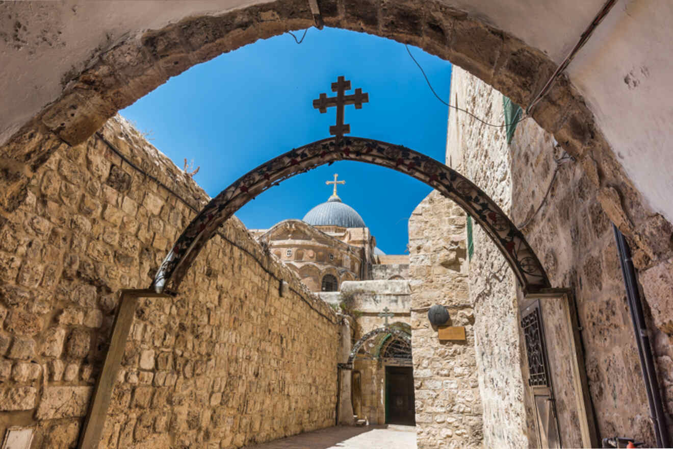 A stone archway topped with two crosses frames a view of an ancient stone church with a domed roof against a clear blue sky.