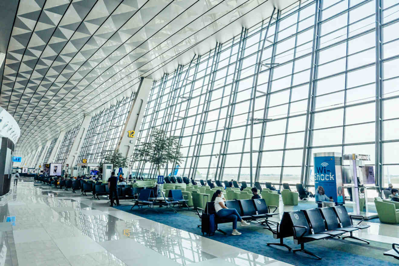 Spacious airport terminal with high ceilings and large windows. Rows of seating areas with some passengers waiting. A Wi-Fi sign and flight gates are visible.