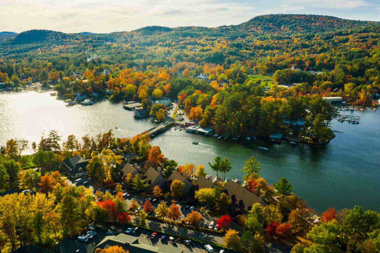 Aerial view of a lakeside town surrounded by vibrant autumn foliage, with houses, docks, and boats visible along the water. Hills with trees extend into the background.