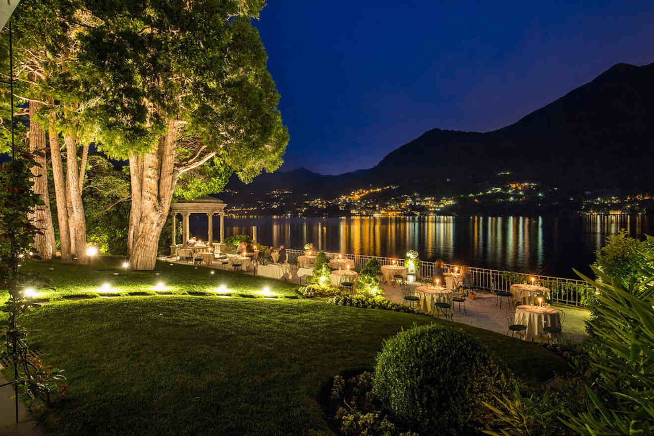 Outdoor dining area by a lake at night, with tables and chairs arranged under trees and a gazebo, illuminated by lights. The lake reflects the lights from nearby hills.